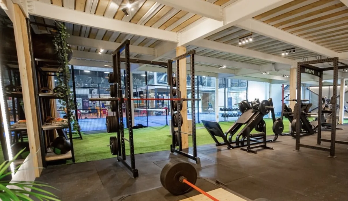Zona de peso libre de Gimnasio Synergy: racks de potencia, barra con discos y las canchas de pádel al fondo